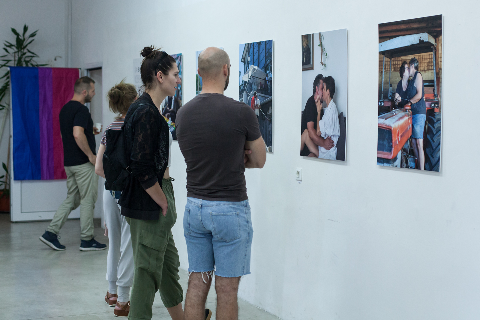 Prizor s otvorenja izložbe "24 BACI" koji pokazuje fotografije queer parova koji se ljube i posjetitelje_ice koji promatraju radove.