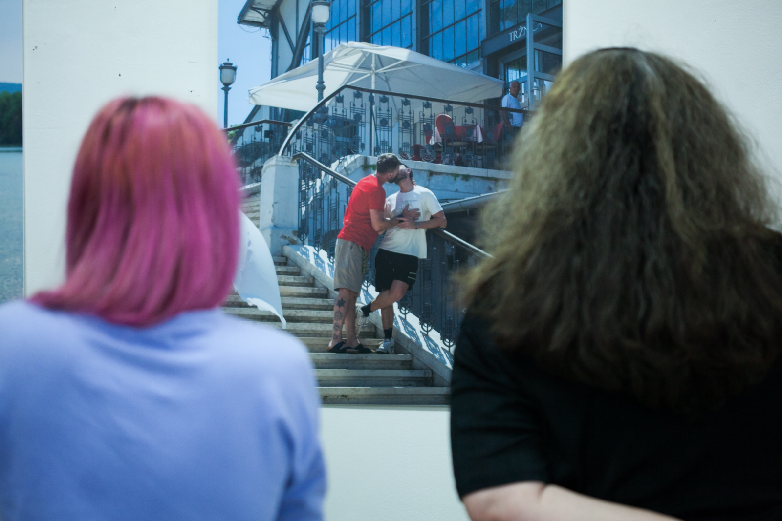 Detalj s izložbe "24 BACI" prikazuje gej par koji se ljubi na stepeništu u javnom prostoru. Iznad njih se nalazi terasa kafića, s koje ih promatra jedna osoba.