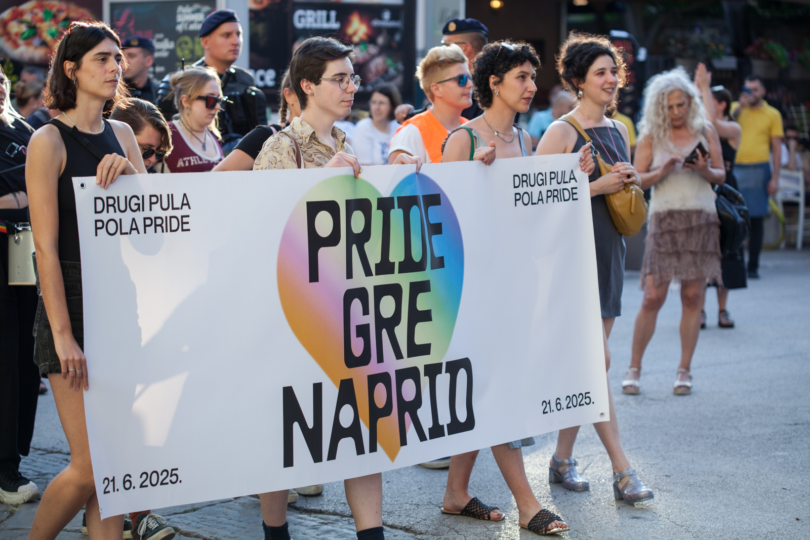 Prizor s drugog pulskog Prajda, koji pokazuje tri osobe koje nose transparent s natpisom "Pride gre naprid", okružene vrevom povorke.