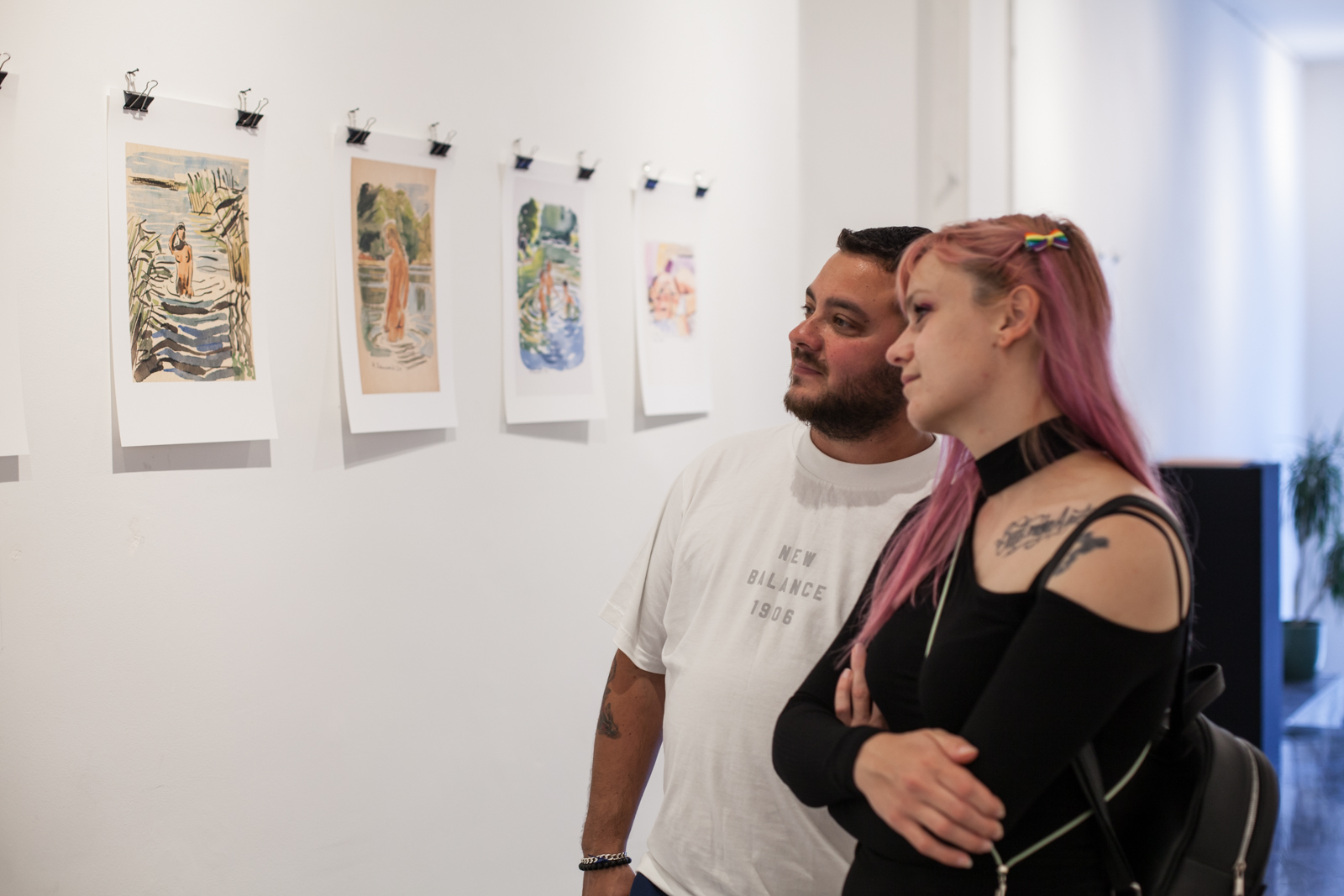 Fotografija prikazuje četiri crteža predstavljena na izložbi "Saphic Sessions", na kojima se gole žene kupaju u prirodi, te posjetitelje_ice koji promatraju crteže.