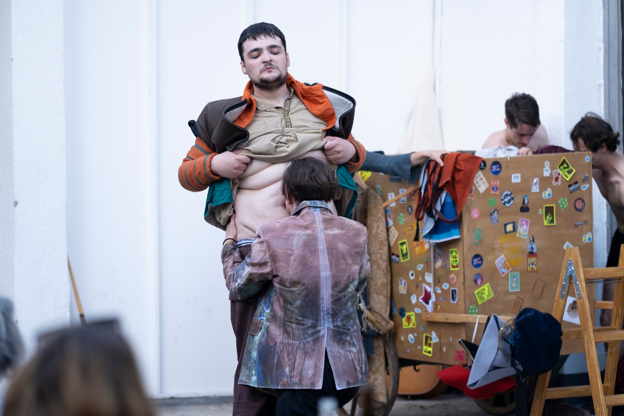 Fotografija prikazuje glumca koji igra Kralja Ubuja kako diže majicu, a pred njime kleči drugi glumac koji ljubi njegov trbuh. U pozadini je pregrada, paravan iza kojega se još dvoje glumaca presvlači.