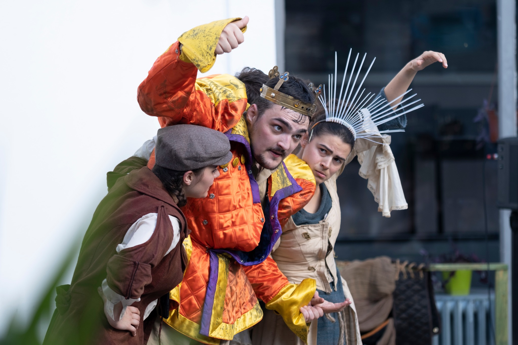Fotografija na kojoj su prikazane dvije glumice namrštenih izraza lica i između njih glumac s kostimom kralja, kojega pridržavaju, odnosno on se na njih oslanja. On je u stiliziranom narančasto-žutom ruhu i s krunom, glumica s njegove lijeve strane nosi krunu od plastike, a glumica s desne strane u kostimu koji ne izgleda kraljevski.