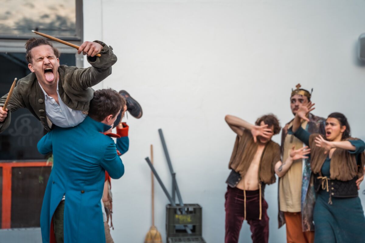 Fotografija prikazuje glumce predstave Ubu Ovo Ono. U prvom planu dva kostimirana glumca drže trećega, koji viče, na ramenima, dok u pozadini vidimo dva glumca i glumicu koji djeluju iznenađeno.