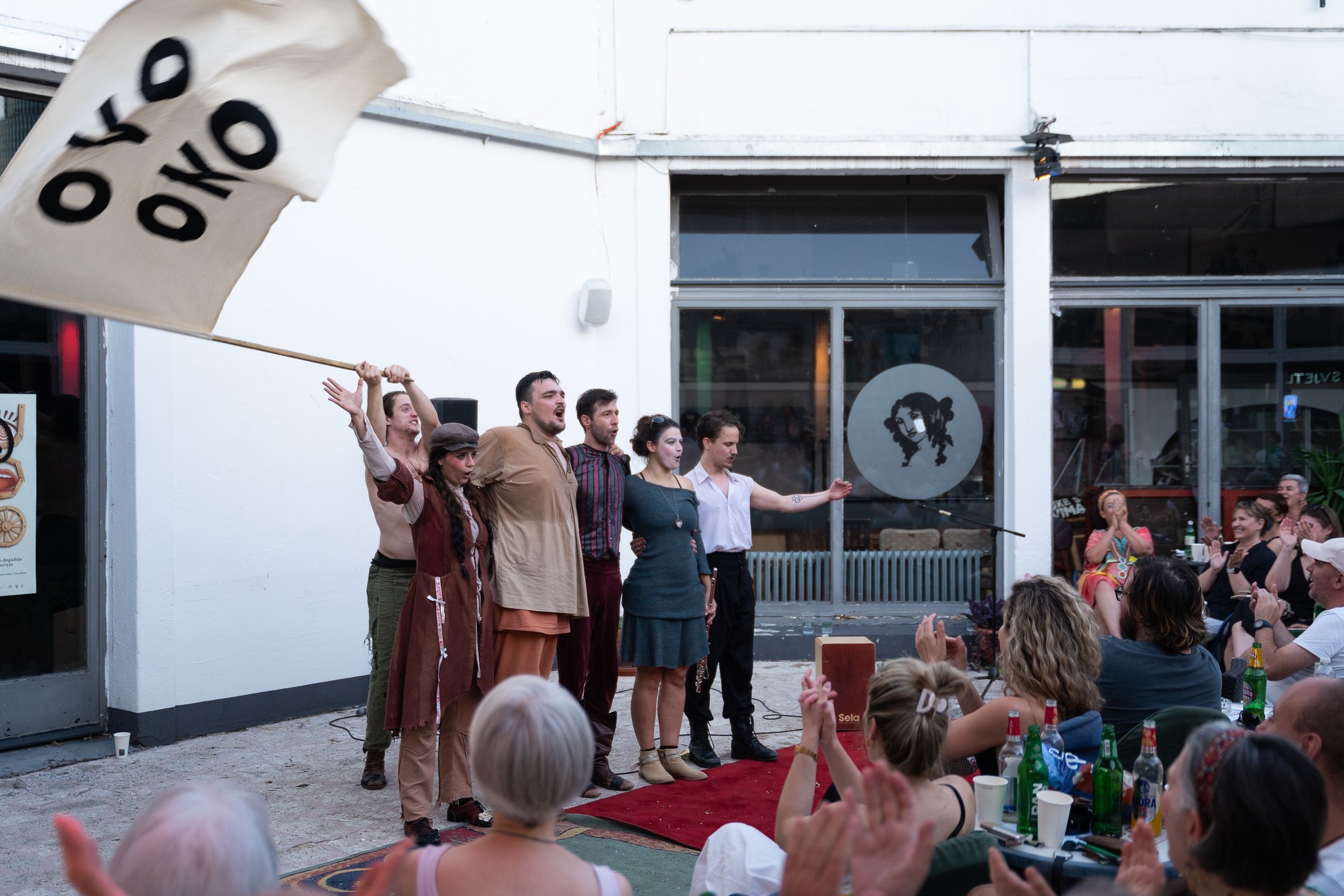 Fotografija prikazuje situaciju glumačkog naklona publici - glumce i publiku koja im plješće. Jedan glumac maše zastavom na kojoj piše "Ovo Ono".